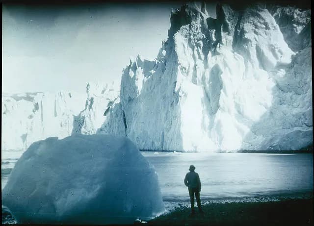 A lone figure stands on a shingle beach before the high ice cliff of a tidewater glacier, with a large ice block in the water.