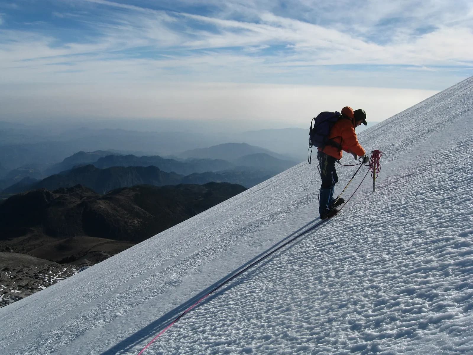 Un alpinista con mochila y cuerda asciende por una pendiente de nieve empinada; al fondo, sierras bajo un mar de nubes.